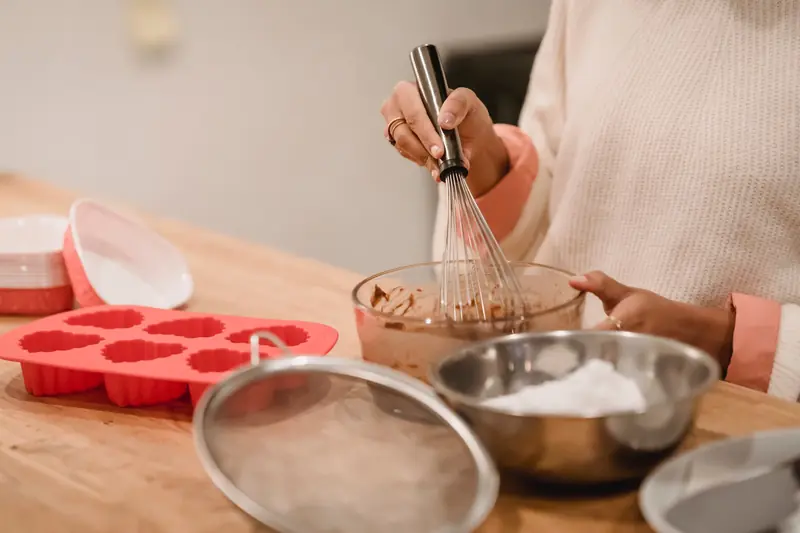 Unrecognizable female baker with whisk mixing ingredients for pastry while standing at table with molds and flour during cooking in kitchen