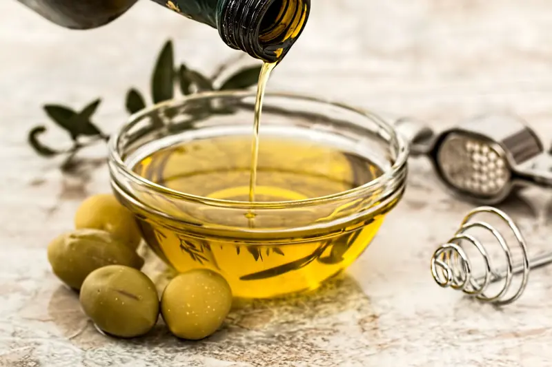 Close-up of olive oil being poured into a glass bowl surrounded by fresh olives and kitchen tools