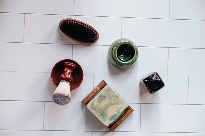 Flat lay of grooming items on white tiles including brush, soap, and jar